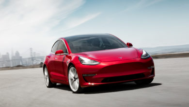 טסלה מודל 3 בצבע אדום על הכביש, מבט מקדמת הרכב כשמאחור עיר ובניינים רחוקים