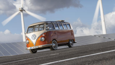 וואן נוסטלגי של פולקסווגן בגרסה מחודשת וחשמלית נוסע על כביש כשמאחוריו טורבינות רוח