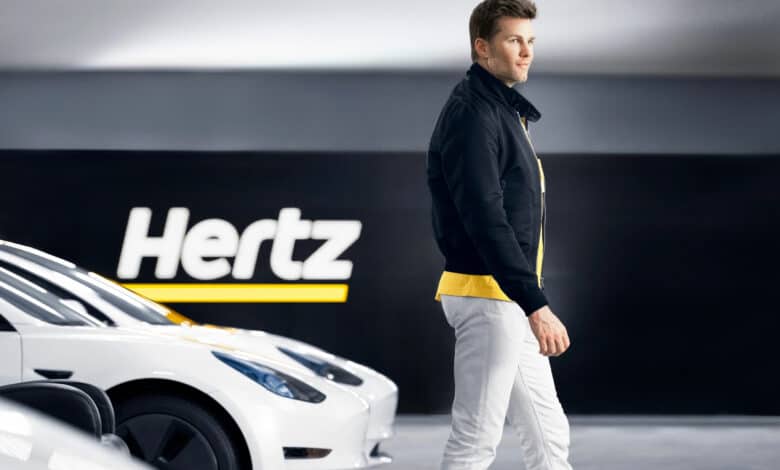 טום בריידי עומד ליד צי של רכבי טסלה מודל 3 עם הלוגו של הרץ מאחוריו