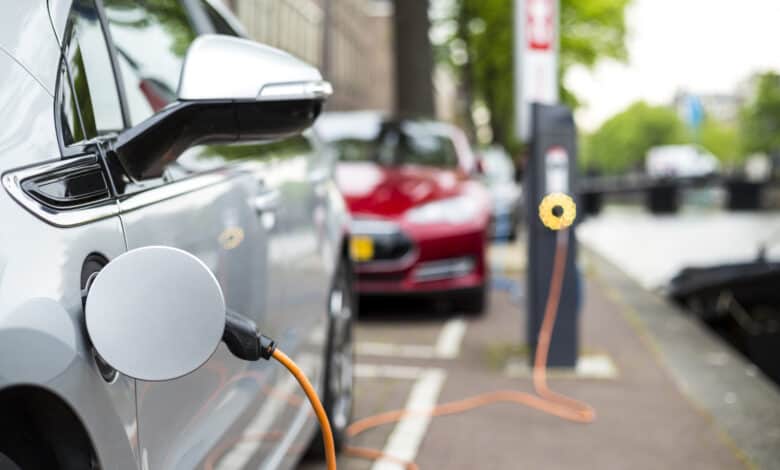 רכב חשמלי לבן מחובר לעמדת טעינה ברחוב ומאחוריו חונה טסלה מודל S אדומה