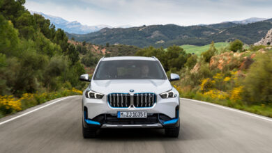 מבט קדמי על ה-BMW iX1 החדשה בצבע לבן עם נגיעות של כחול, עם גריל גדול מקדימה בנסיעה מהירה על כביש