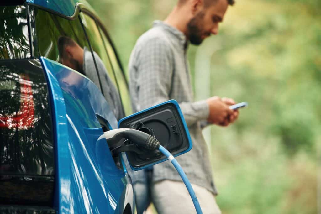 אדם נשען על רכב חשמלי ומשתמש בפלאפון בזמן שהרכב נטען